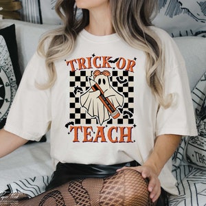 Op de afbeelding: Wit T-shirt met een zwart-wit geruite achtergrond. Een geest met een rode bandana houdt een potlood vast met de tekst "Teach Like a Boss" erop. De tekst "Trick or Teach" is geschreven in oranje en zwarte letters.