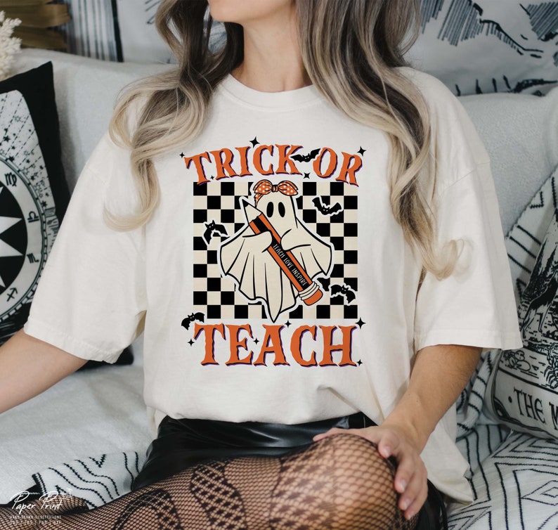 Op de afbeelding: Wit T-shirt met een zwart-wit geruite achtergrond. Een geest met een rode bandana houdt een potlood vast met de tekst "Teach Like a Boss" erop. De tekst "Trick or Teach" is geschreven in oranje en zwarte letters.