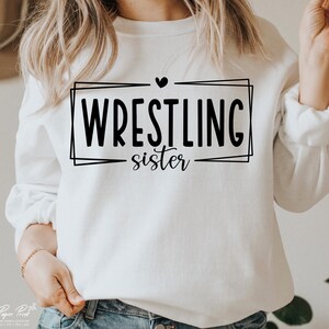 May include: White sweatshirt with black text that reads "Wrestling sister" inside a black frame with a small black heart above the text.