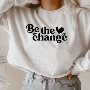 Puede incluir: Una sudadera gris con el texto "Be the change" en negro con un s&iacute;mbolo de coraz&oacute;n.