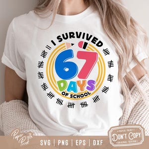 May include: White t-shirt with a colorful graphic that reads "I SURVIVED 67 DAYS OF SCHOOL." The design includes a blue "6", a pink "7", and the word "DAYS" in green and purple. The shirt has a round neck and short sleeves.