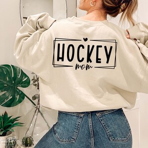 以下が含まれることがあります： クリーム色のスウェットシャツに、黒いグラフィックで「HOCKEY mom」と箱の中に書かれており、テキストの上に小さなハートがあります。