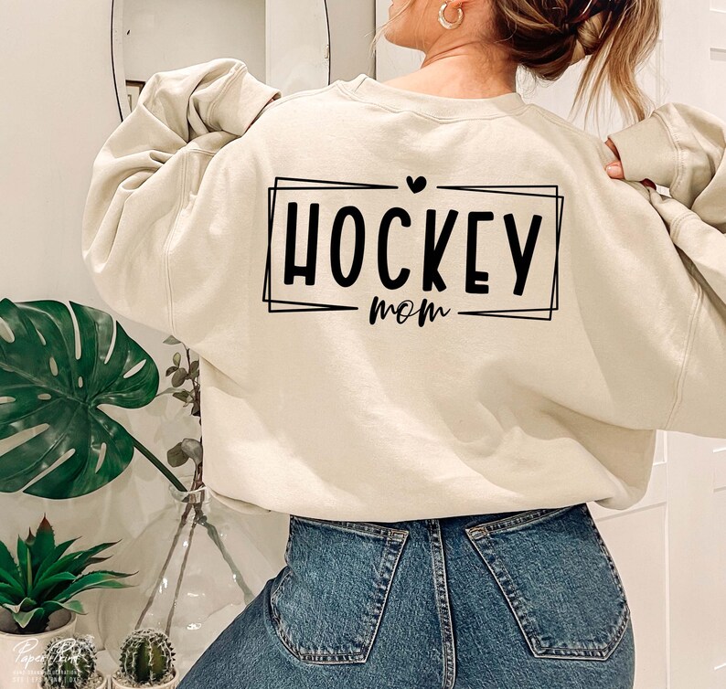 以下が含まれることがあります： クリーム色のスウェットシャツに、黒いグラフィックで「HOCKEY mom」と箱の中に書かれており、テキストの上に小さなハートがあります。