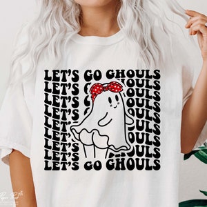 May include: White t-shirt featuring a black and white graphic of a ghost wearing a red bandana with white stars. The text "LET'S GO GHOULS" is repeated in a retro font.