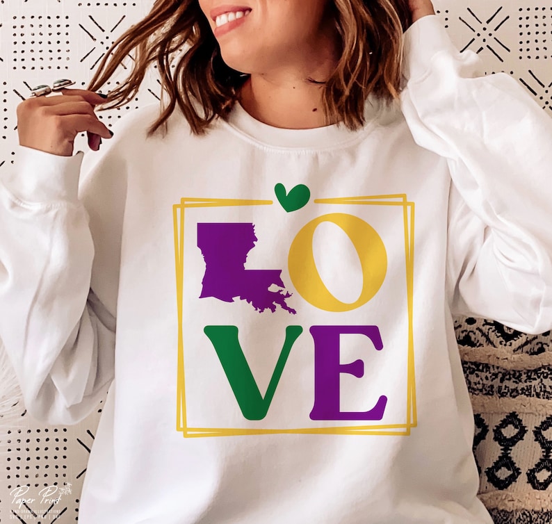 May include: White sweatshirt with a graphic design of the word "LOVE" in green, yellow, purple, and white. The letter "O" is a circle and the letter "L" is the outline of the state of Louisiana. A small green heart is above the word.