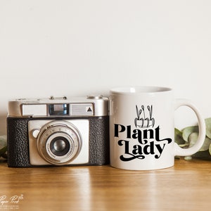 May include: White ceramic mug with a black graphic that says "Plant Lady" and a small illustration of a potted plant. The mug is sitting on a wooden surface next to a vintage camera.