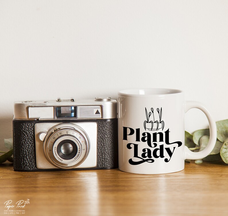 May include: White ceramic mug with a black graphic that says "Plant Lady" and a small illustration of a potted plant. The mug is sitting on a wooden surface next to a vintage camera.