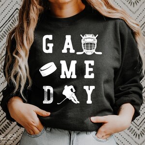 K&ouml;nnte beinhalten: Schwarzer Pullover mit wei&szlig;em Text, der "GAME DAY" lautet, mit einem Hockeyschl&auml;ger, einem Puck und einem Hockeyhelm.