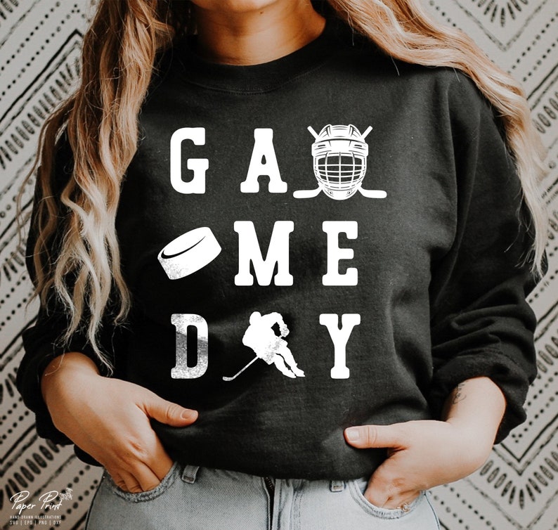 K&ouml;nnte beinhalten: Schwarzer Pullover mit wei&szlig;em Text, der "GAME DAY" lautet, mit einem Hockeyschl&auml;ger, einem Puck und einem Hockeyhelm.