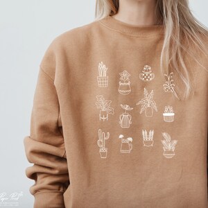 May include: A brown sweatshirt with a white outline design of various potted plants. The design includes cacti, succulents, and leafy plants.