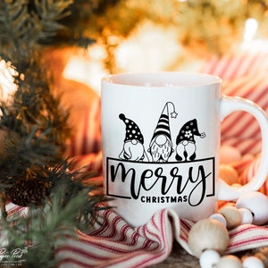 K&ouml;nnte beinhalten: Wei&szlig;e Keramiktasse mit einem schwarzen Design aus drei Gnomen mit dem Text "merry christmas".