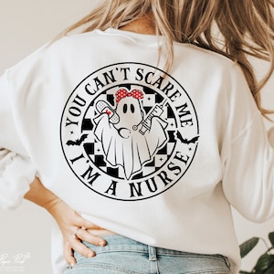 Puede incluir: Sudadera blanca con un gr&aacute;fico en blanco y negro de un fantasma con un pa&ntilde;uelo rojo, sosteniendo un vendaje y una jeringa. El gr&aacute;fico est&aacute; dentro de un c&iacute;rculo con el texto "You Can't Scare Me I'm A Nurse".