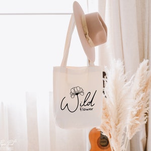 May include: A white canvas tote bag with black text that reads "Wild Flower" and a stylized flower graphic. The bag is hanging on a white rack with a straw hat hanging on the rack above the bag.