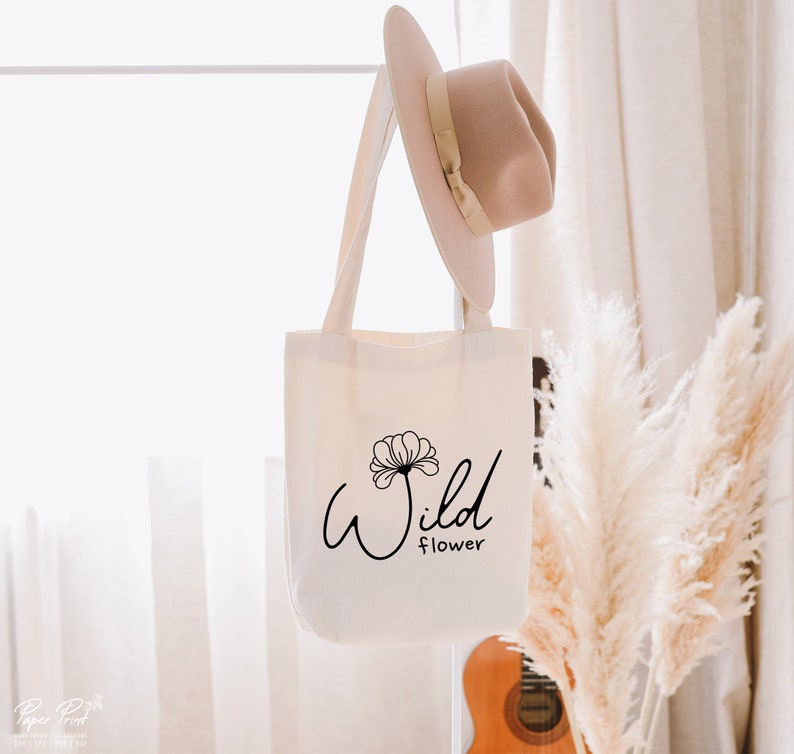 May include: A white canvas tote bag with black text that reads "Wild Flower" and a stylized flower graphic. The bag is hanging on a white rack with a straw hat hanging on the rack above the bag.