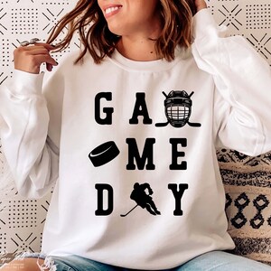 K&ouml;nnte beinhalten: Wei&szlig;er Pullover mit schwarzem Text, der "GAME DAY" lautet, mit einem Grafikmotiv aus Hockeyschl&auml;ger, Puck und Helm.