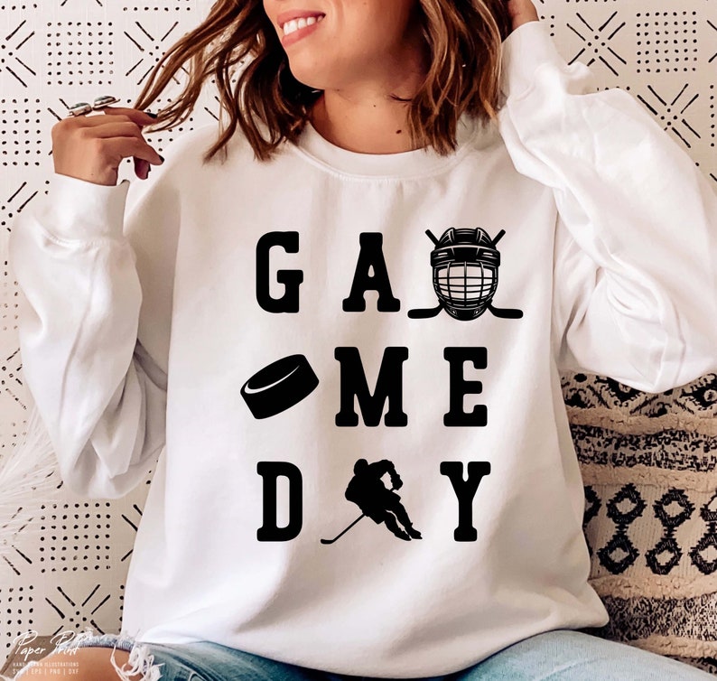 K&ouml;nnte beinhalten: Wei&szlig;er Pullover mit schwarzem Text, der "GAME DAY" lautet, mit einem Grafikmotiv aus Hockeyschl&auml;ger, Puck und Helm.