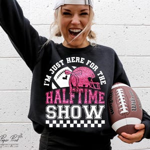Può includere: Felpa nera con testo bianco che recita "I'm just here for the halftime show". Il testo è circondato da un pallone da calcio bianco e un casco da football rosa glitterato. Un pallone da calcio marrone e bianco è tenuto nella mano destra della persona che indossa la felpa.