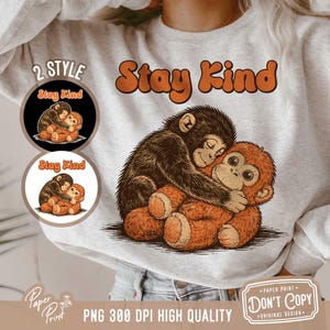 以下が含まれることがあります： オレンジ色の文字で「Stay Kind」と書かれた、明るい灰色のスウェットシャツ。スウェットシャツには、抱き合っている2匹の漫画の猿のグラフィックが描かれています。2つの小さな円形のグラフィックは、同じ猿のデザインを示しています。