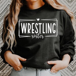 May include: Black sweatshirt with white text that reads "WRESTLING sister" inside a double-lined rectangle with a small heart above the text.