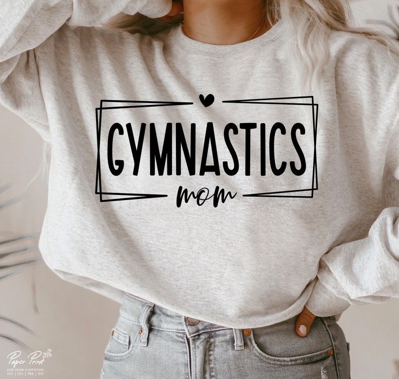 May include: A light gray sweatshirt with the words "GYMNASTICS mom" in black script inside a black rectangular frame. A small black heart is above the word "GYMNASTICS". The sweatshirt is made of a soft material.