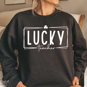 Puede incluir: Sudadera negra con un dise&ntilde;o gr&aacute;fico blanco que dice "LUCKY teacher" con un tr&eacute;bol por encima de la palabra "LUCKY".