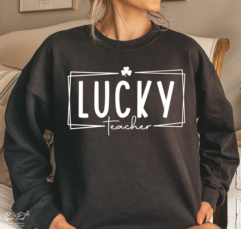Puede incluir: Sudadera negra con un dise&ntilde;o gr&aacute;fico blanco que dice "LUCKY teacher" con un tr&eacute;bol por encima de la palabra "LUCKY".