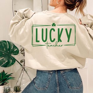 Puede incluir: Una sudadera de color crema con el texto "LUCKY teacher" en letras verdes dentro de un cuadro verde con un tr&eacute;bol por encima del texto.