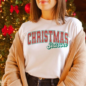 May include: A white t-shirt with a red and green vintage-style graphic that says "Christmas Season".