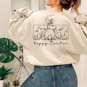 May include: A cream-coloured sweatshirt with a black and white illustration of a bunny rabbit in a field of flowers. The text "happy Easter" is printed below the illustration.