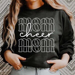May include: Black sweatshirt with the words "mom cheer mom" in white block letters. The word "cheer" is in a cursive font. The sweatshirt is a crew neck style. The background is a black and white patterned design.