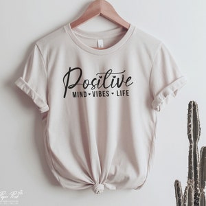 May include: A beige t-shirt with the text "Positive Mind - Vibes - Life" printed in black.
