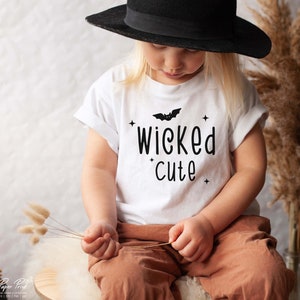 May include: A white t-shirt with a black bat and the text "wicked cute" printed on it. A child is wearing the shirt and a black hat.