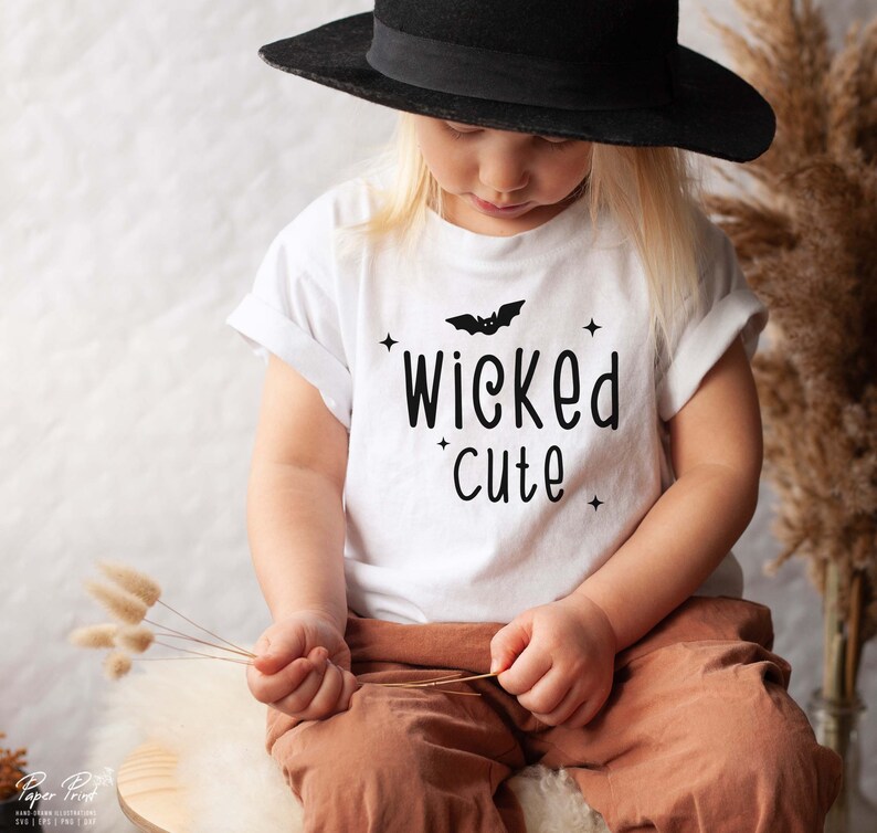 May include: A white t-shirt with a black bat and the text "wicked cute" printed on it. A child is wearing the shirt and a black hat.