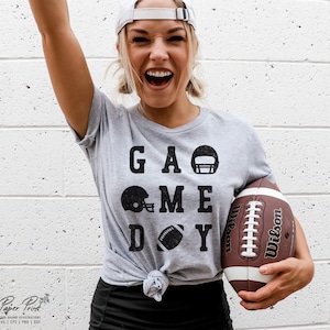 May include: A gray t-shirt with the text "GAME DAY" in black letters. The text is surrounded by black football helmet and football graphics. A person is wearing the shirt and holding a brown football.