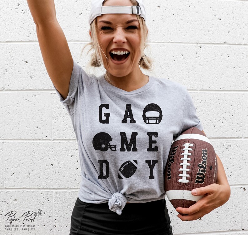 May include: A gray t-shirt with the text "GAME DAY" in black letters. The text is surrounded by black football helmet and football graphics. A person is wearing the shirt and holding a brown football.