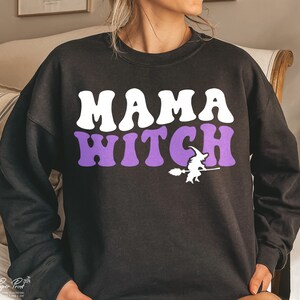 Pu&ograve; includere: Felpa nera con il testo "MAMA WITCH" in lettere bianche e viola. Sotto il testo c'&egrave; una piccola silhouette di una strega che vola su una scopa.