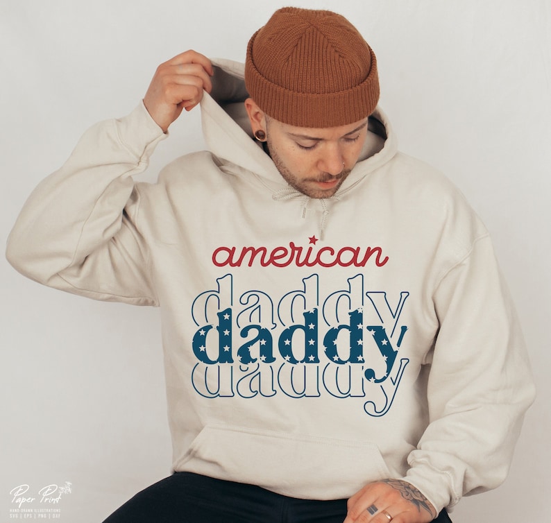 Puede incluir: Una sudadera con capucha beige con el texto "american daddy" en rojo y azul con un patr&oacute;n de estrellas. El texto est&aacute; apilado sobre s&iacute; mismo con la capa superior en rojo y las dos capas inferiores en azul con estrellas blancas.