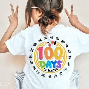May include: White t-shirt with a colorful graphic that says "100 Days of School". The graphic features a rainbow-colored circle with the number 100 in the center. There are black hash marks around the circle.