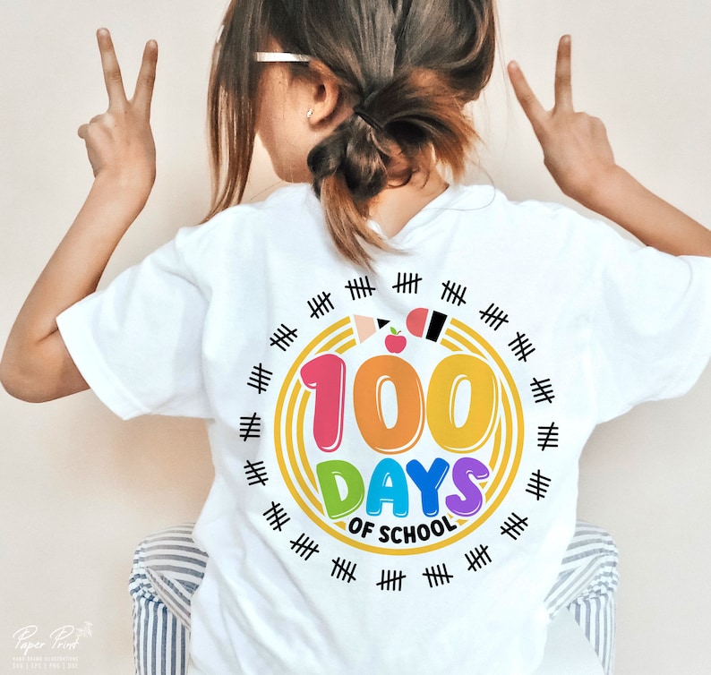 May include: White t-shirt with a colorful graphic that says "100 Days of School". The graphic features a rainbow-colored circle with the number 100 in the center. There are black hash marks around the circle.