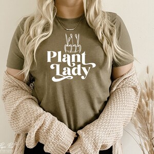 May include: A woman wearing a green t-shirt with the text "Plant Lady" in white lettering. The shirt features a simple line drawing of a potted plant with leaves.