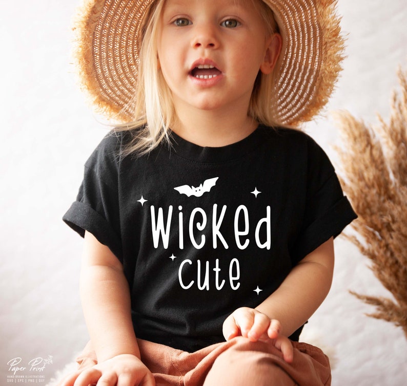 May include: A black t-shirt with a white bat and the text "wicked cute" on it. A young child is wearing the shirt and a straw hat.
