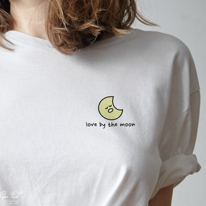 May include: White t-shirt with a yellow crescent moon graphic and the text "love by the moon".