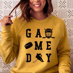 K&ouml;nnte beinhalten: Ein gelber Pullover mit dem Text "GAME DAY" in Schwarz. Der Text ist von einem Hockeyschl&auml;ger, einem Puck und einem Hockeyhelm umgeben.