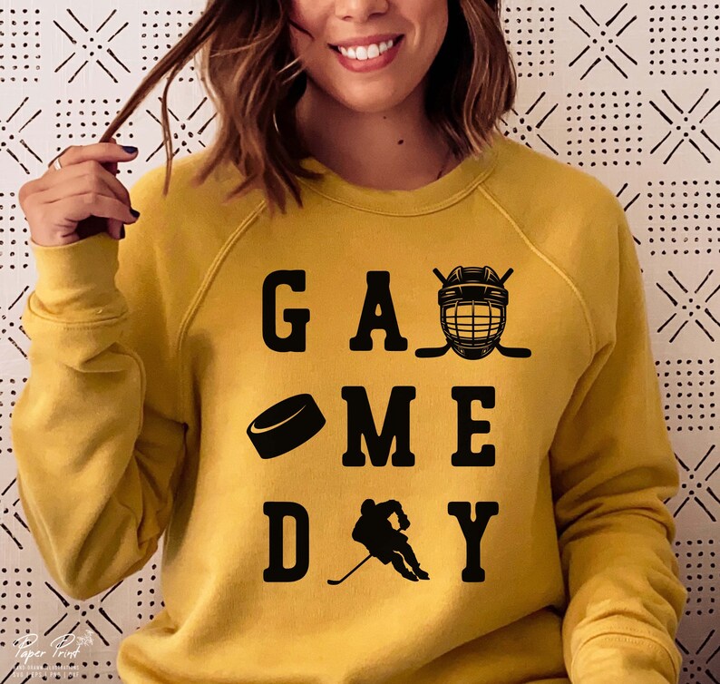 K&ouml;nnte beinhalten: Ein gelber Pullover mit dem Text "GAME DAY" in Schwarz. Der Text ist von einem Hockeyschl&auml;ger, einem Puck und einem Hockeyhelm umgeben.