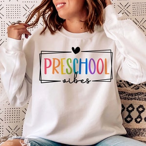 May include: White sweatshirt with a rainbow colored "PRESCHOOL vibes" graphic and a black heart.