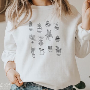 May include: White sweatshirt with a black line drawing of various potted plants. The plants include cacti, succulents, and leafy plants.