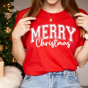 K&ouml;nnte beinhalten: Ein rotes T-Shirt mit wei&szlig;em Text, der "Merry Christmas" in einer Vintage-Universit&auml;ts-Schriftart lautet.