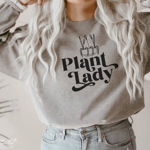 May include: A gray sweatshirt with the text "Plant Lady" in black script font. A black and white illustration of a potted plant with leaves is above the text.
