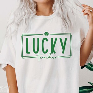 Puede incluir: Camiseta blanca con un tr&eacute;bol verde y el texto "LUCKY teacher" en un cuadro verde.