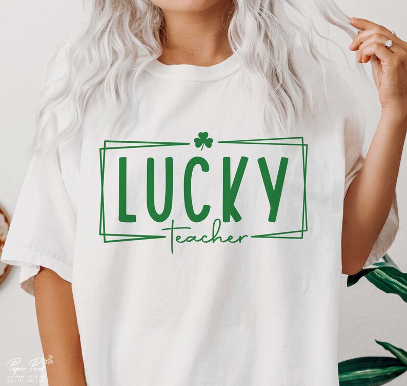 Puede incluir: Camiseta blanca con un tr&eacute;bol verde y el texto "LUCKY teacher" en un cuadro verde.